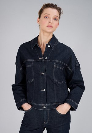 Frau mit den Händen in den Taschen, trägt eine dunkelblaue Jeansjacke mit silbernen Knöpfen und passende Jeans vor einem schlichten grauen Hintergrund.