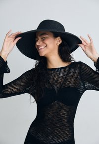 Copertura in crochet nera con maniche lunghe, motivo testurizzato e scollatura profonda. Abbinata a un cappello di paglia nera a tesa larga.