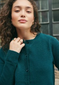 Žena s vlnitými vlasy má na sobě tyrkysový cardigan na knoflíky, zlaté náušnice a dva zlaté prsteny, z nichž jeden má zelený kámen, stojí blízko skleněných oken.