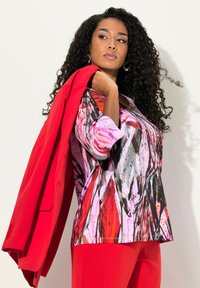 Multifarvet bluse med pink, rød og sort abstrakt print, båret med en rød blazer. Sammen med matchende røde bukser. Krøllet sort hår.