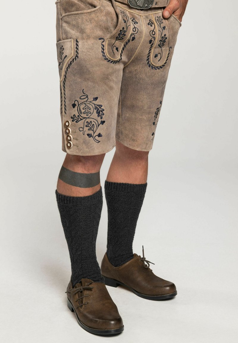 Braune Shorts mit aufwändiger blauer Stickerei, seitlichem Knopfdetail, kombiniert mit dunklen gestrickten Socken und braunen Schuhen.