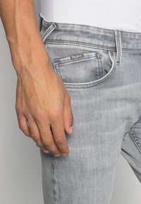 Vaqueros de denim gris con un acabado desgastado, que cuentan con un bolsillo frontal y una etiqueta de marca visible en la cintura. Mano descansando sobre el muslo.