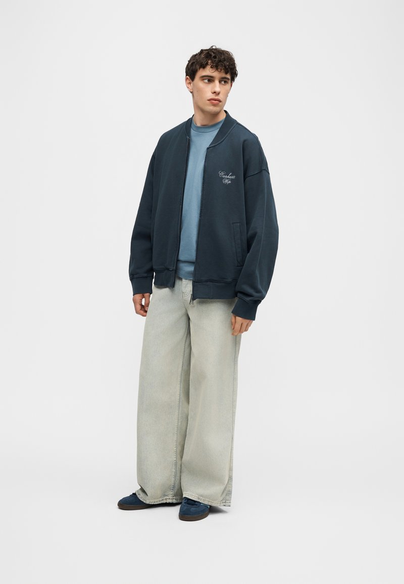 Young man wearing dark navy bomber jacket, light blue sweatshirt, wide light gray jeans, and navy sneakers, standing and looking to the side.