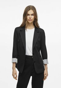 Černý blazer se strukturovaným designem, zapínáním na jeden knoflík a ohrnutými pruhovanými manžetami. Nosený přes bílou košili a černé kalhoty.
