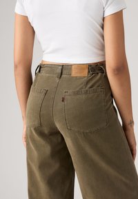 Personne portant un jean Levi's vert olive avec des poches arrière et un t-shirt blanc court, visible de la taille vers le bas.
