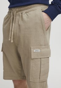 Pantalones cortos de carga beige en una tela suave y texturizada, con bolsillos laterales y una etiqueta en la pierna derecha. Cintura elástica con detalles de cordón.