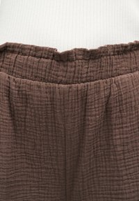 Pantalon marron avec taille élastique texturée sur fond blanc, montrant les détails du tissu et la taille froncée.