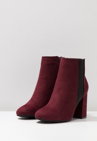 Par de botines tobilleros de ante burdeos con tacones de bloque y cremalleras laterales, exhibidos sobre una superficie blanca con un fondo gris liso.