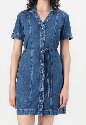 Kvinde iført en kortærmet blå denim kjole med knapper, krave og bindebånd i taljen mod en ensfarvet baggrund.