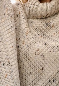 Knitted sweater in a textured cream color with flecks of brown and gray. Features a thick, ribbed turtleneck and wide sleeves.
