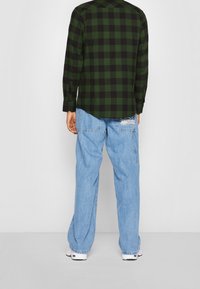 Camicia a maniche lunghe a quadri verdi e neri con orlo arrotondato, abbinata a jeans a gamba larga di colore azzurro chiaro con tasche laterali e logo sul retro.