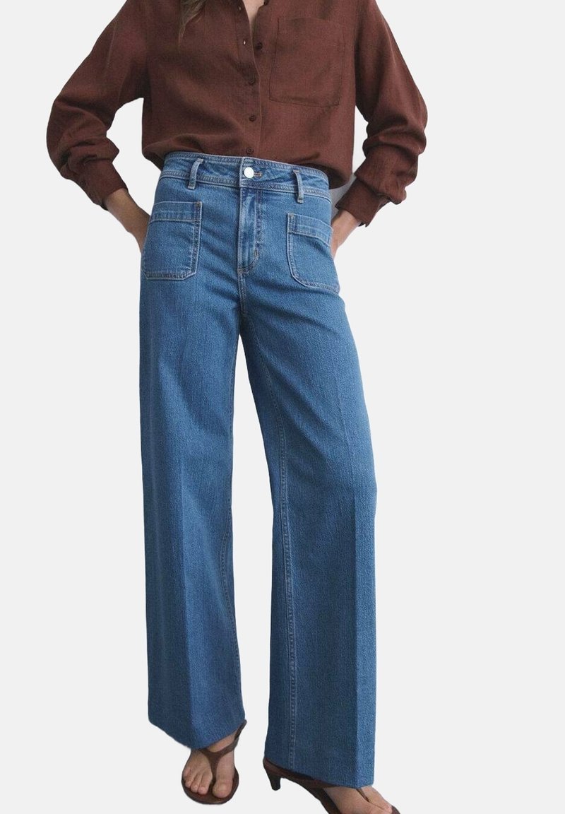 Jean en denim bleu à jambes larges avec une taille haute, doté de deux poches avant et d'une fermeture à boutons. Associé à une chemise marron à manches longues.