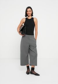 Haut noir sans manches, pantalon gris à jambes larges, chaussures plates noires et pochette noire. Textures simples et lignes épurées partout.