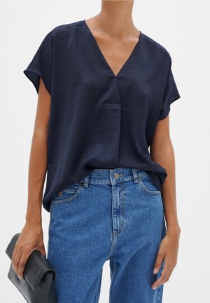 Person iført en løs mørkeblå V-hals kortærmet bluse, der er stoppet ned i højttaljede blå jeans, og holder en sammenfoldet sort clutch.