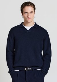 Navyblauer gerippter Strickpullover mit Überschlagkragen, langen Ärmeln und lockerer Passform. Wird über einem weißen Rundhalsausschnitt-Shirt getragen, kombiniert mit dunklen Hosen.