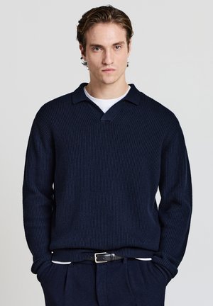 Pullover in maglia a coste blu navy con colletto incrociato, maniche lunghe e vestibilità rilassata. Indossato sopra una t-shirt bianca a girocollo, abbinato a pantaloni scuri.