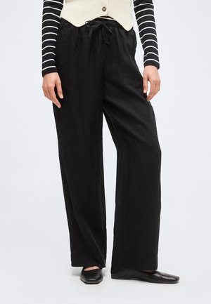 Femme portant un pantalon noir ample à cordon, des ballerines noires, un haut à manches longues rayé noir et blanc, et un gilet crème boutonné.