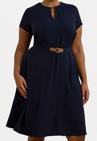 Vestido navy con escote en forma de llave, mangas cortas y una cintura ajustada acentuada por un cinturón marrón. La tela suave crea una silueta de falda acampanada.