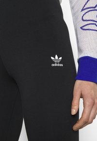 Černé legíny vyrobené z elastické látky, s bílým logem Adidas na stehně. Detailní záběr ruky proti látce.