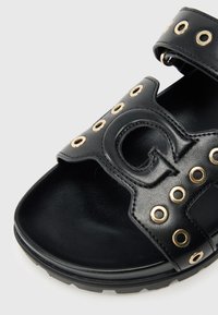 Μαύρο δερμάτινο σανδάλι με χρυσαφί μεταλλικά κορδόνια, εμφανές κόψιμο με το λογότυπο και υφασμένο πέλμα. Ρυθμιζόμενο λουράκι για σταθερή εφαρμογή.