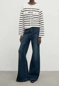 Kragenloser weißer Pullover mit schwarzen Streifen und zwei Brusttaschen; kombiniert mit weit geschnittenen blauen Jeans im Denim-Stil mit ausgefranstem Saum.