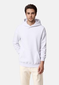 Hoodie blanc en tissu doux, avec une poche frontale, une capuche à cordon de serrage et des poignets côtelés. Associé à un pantalon de couleur claire.