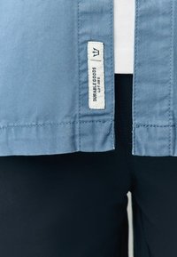 Šviesiai mėlyna audinio marškiniai su tiesiu kraštu, su baltu etiketu, ant kurio užrašyta "Durable Goods Lot 02", esantis arti apačios. Matosi juodos kelnės.