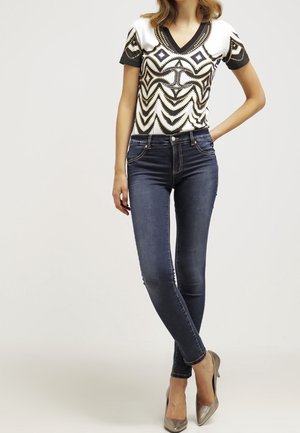 Kobieta ubrana w białą i czarną, wzorzystą bluzkę z krótkim rękawem, ciemnoniebieskie obcisłe jeansy oraz metaliczne czółenka na wysokim obcasie z noskiem w szpic, stojąca z jedną ręką na biodrze.