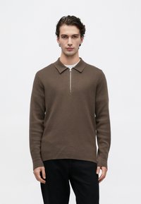 Un joven de pie, erguido, vistiendo un suéter de punto marrón de manga larga con un cuello con cremallera en la parte delantera y pantalones negros, contra un fondo blanco simple.