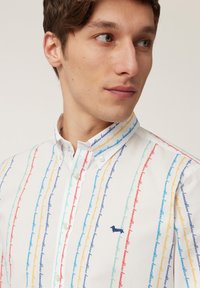 Witte shirt met multikleurige verticale lijnen in blauw, rood, geel en groen, met een klein blauw geborduurd hondenlogo op de borst.