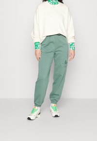 Ljusgröna sweatpants med elastiska muddar, med en diskret logotyp vid höften. Matchas med en vit sweatshirts och ett mönstrat grönt halslager.