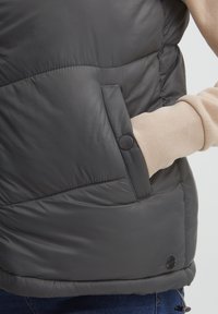 Tamno siva puffer prsluk s glatkom, sjajnom teksturom, sa kutnim šavovima i bočnim džepom s kopčom na dugme.
