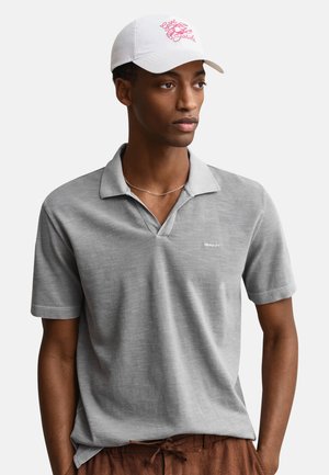 Homme portant un polo gris clair, une casquette blanche avec un logo rose de crabe, un pantalon marron à cordon et un collier fin en argent, regardant sur le côté.