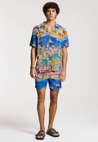 Short-sleeve button-up shirt and matching shorts in blue fabric with colorful coastal patterns, featuring boats and palm trees. Sandals worn.