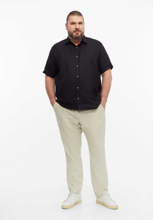 Homme barbu debout, les mains dans les poches, portant une chemise noire à manches courtes, un pantalon beige et des baskets blanches sur fond blanc.