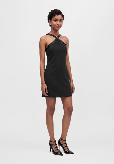 Une femme porte une mini-robe noire à bretelles croisées et des chaussures noires à talons hauts sur un fond clair et uni.