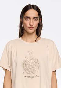 T-shirt beige a maniche corte con un design stampato di un ananas con fiori, testo che recita "BimBaY LoLa" e un tessuto testurizzato.