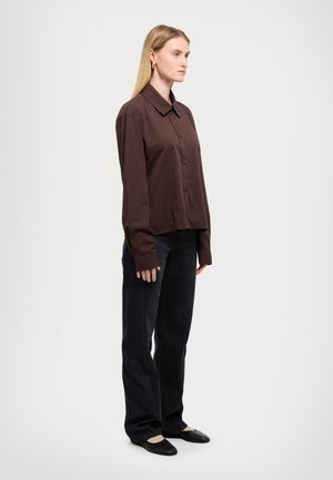 Brun knappopp skjorte med lange ermer, parret med svarte vide jeans og svarte leiligheter, tatt fra siden mot en ensfarget bakgrunn.