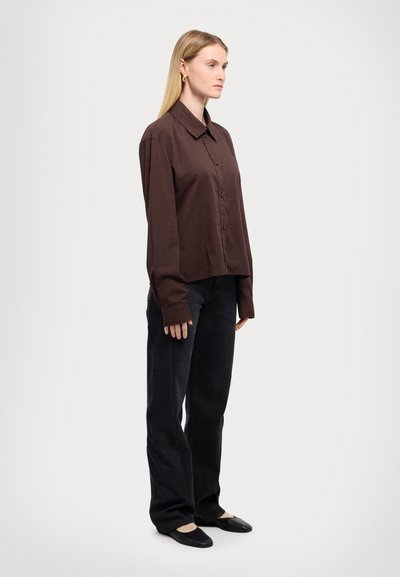 Chemise boutonnée marron à manches longues, portée avec un jean noir à jambes larges et des ballerines noires, photographiée de côté sur un fond uni.