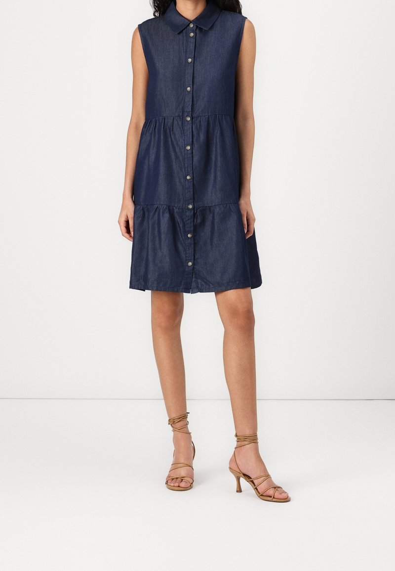 Robe bleu foncé sans manches à boutons sur le devant avec col, portée avec des sandales à talons à brides couleur camel, mannequin posant sur fond blanc.
