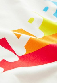 Valge T-särk, millel on suur, värvikas gradienttekst punases, oranžis, kollases, rohelises ja sinises. Pehme kangastekstuuri ja siledaga pind.