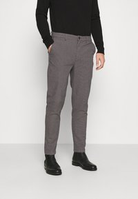 Man med slim-fit grå rutiga byxor, svart långärmad skjorta och svarta läderankelstövlar, stående mot en enkel bakgrund.