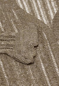 Világosbarna kötött ruhadarab texturált mintával; nyitott varratokkal és hullámos mintákkal rendelkezik, lágy, fémes fényű megjelenést kölcsönözve.