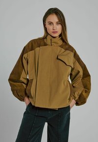 Braune Jacke aus weichem Stoff mit hohem Kragen, versehen mit gerippten Schulterakzenten, einer Fronttasche und elastischen Bündchen.