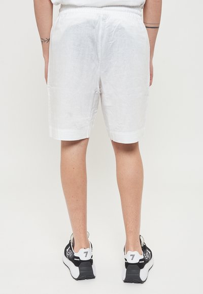 Pantaloni corti bianchi in lino con vita elastica, dal taglio casual, con risvolti e indossati con scarpe da ginnastica bianche e nere.