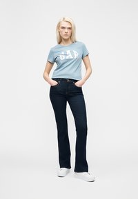 Jeune femme debout les mains dans les poches, portant un t-shirt GAP bleu clair, un jean bleu foncé et des baskets blanches à plateforme sur fond blanc.