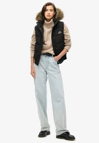 Schwarze Pufferweste mit einem mit Fell gefütterten Kapuzen, getragen über einem beigen Rollkragenpullover. Hellblaue Schlagjeans und schwarze Schuhe runden das Outfit ab.