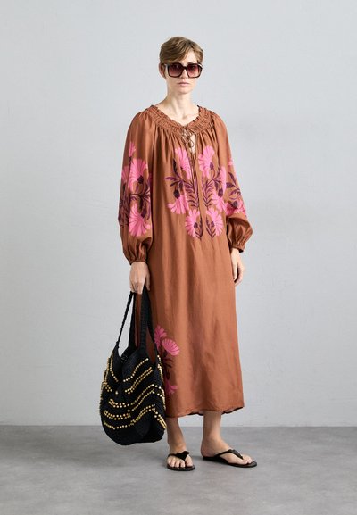 Bruine bloemenjurk met roze en paarse patronen, lange mouwen en een losse pasvorm. Zwarte geweven tas met gouden accenten. Zwarte flip-flops.