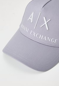 Šviesiai violetinė kepurė, pagaminta iš tekstūruoto audinio, su balta išsiuvinėta logo "A|X" ir "Armani Exchange" priekinėje juostoje. Išlenktas kraštas.