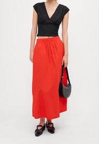 Sort cropped top med korte ærmer, parret med en lang, svajende rød nederdel. Sort taske med hvide nitte-detaljer og sorte sko.